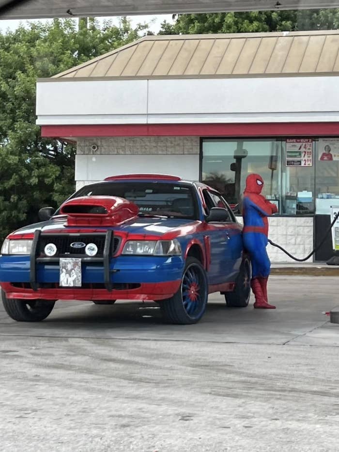 A person dressed as Spider-Man is fueling a customized car at a gas station. The car is styled with red and blue details