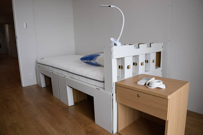 A minimalistic room with a white single bed, a wooden nightstand, a flexible reading lamp attached to the bed, and a remote control on the nightstand
