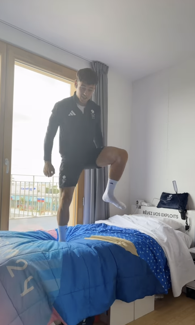 An athlete in athletic wear poses mid-step on a bed with "REUZ UOS EXPLOITS" printed on the headboard