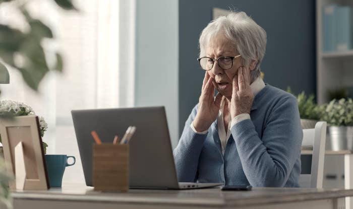 Elderly woman with glasses and a blue sweater sits at a desk, looking surprised at her laptop screen, hands on her cheeks. Office setting in the background