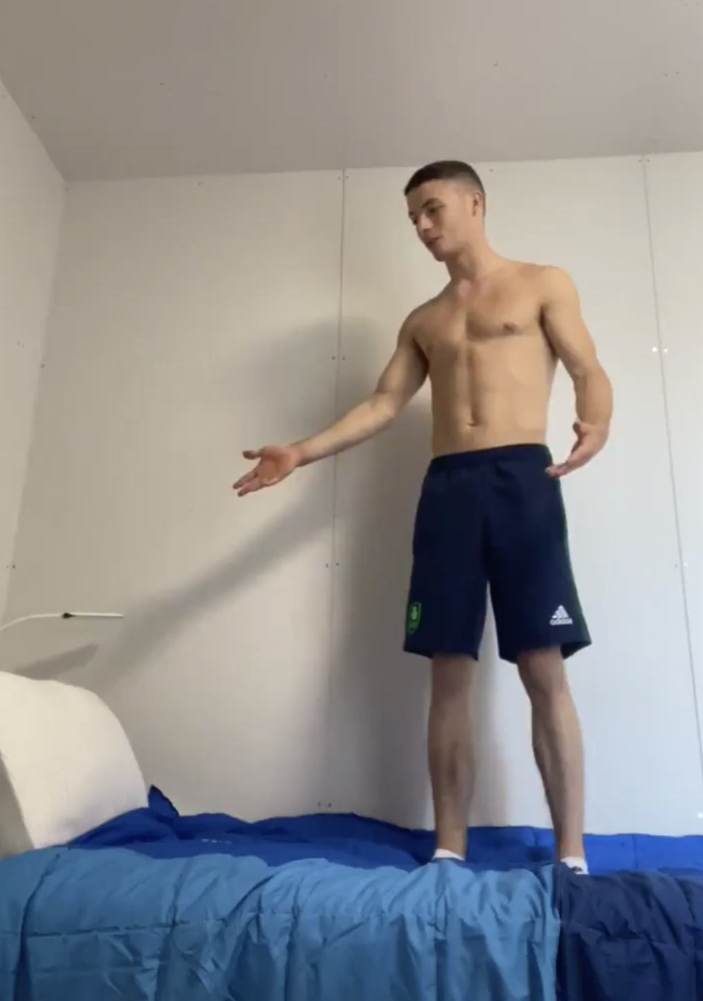 A shirtless man stands on a bed wearing navy blue shorts, extending one arm as if gesturing towards something off-camera