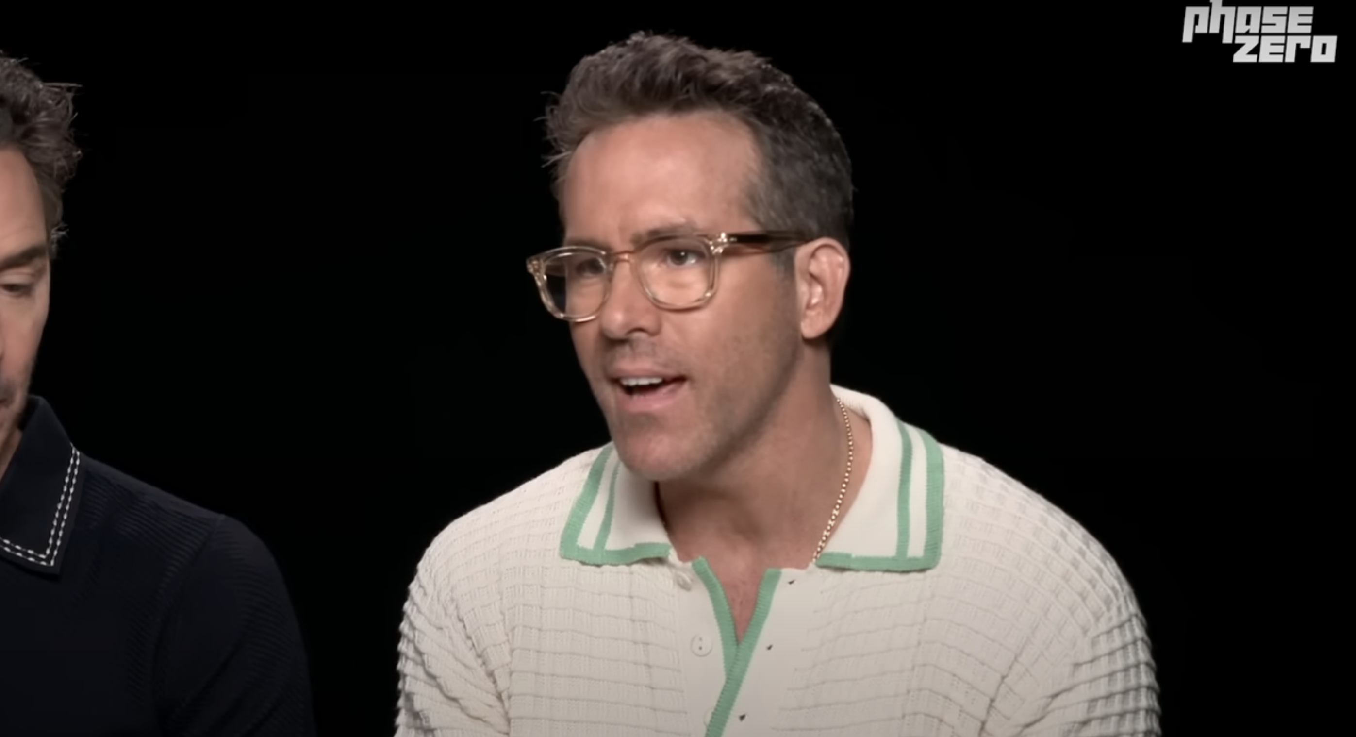 Ryan Reynolds in a casual white textured shirt with green trim and clear glasses during an interview. Another person partially visible on the left