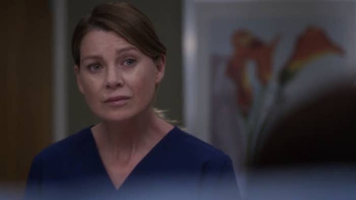 Meredith Grey, dressed in blue medical scrubs, looking concerned, stands in what appears to be a hospital setting
