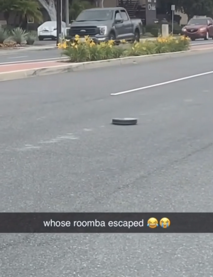A Roomba vacuum is seen in the middle of a street. Text on the image reads, "whose roomba escaped ??"