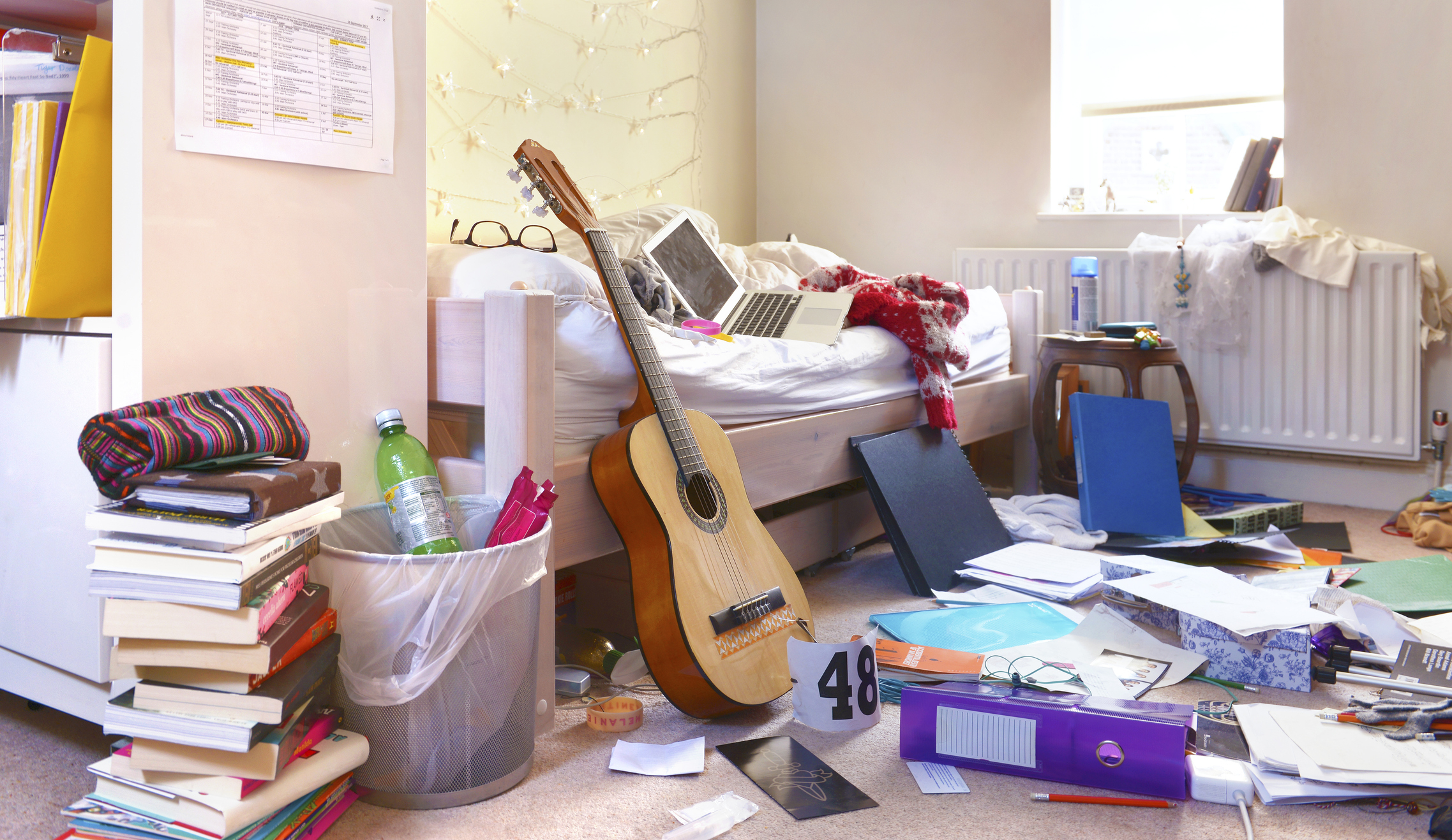 A cluttered bedroom with books, a guitar, a laptop on the bed, a trash bin holding a bottle, and various papers strewn across the floor