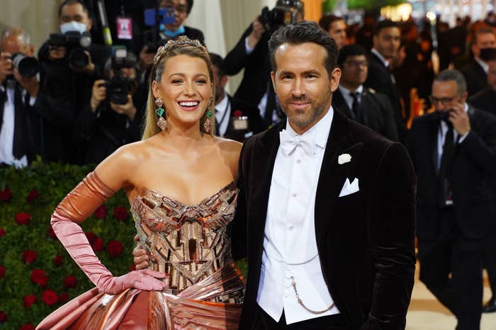 Blake Lively in a strapless gown and Ryan Reynolds in a tuxedo pose together on the red carpet, smiling at the camera. Photographers are in the background
