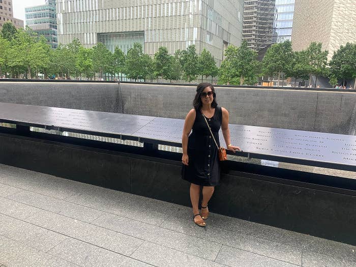 Reviewer posing in front of a memorial structure with engraved names, standing on a pavement with modern buildings and trees in the background