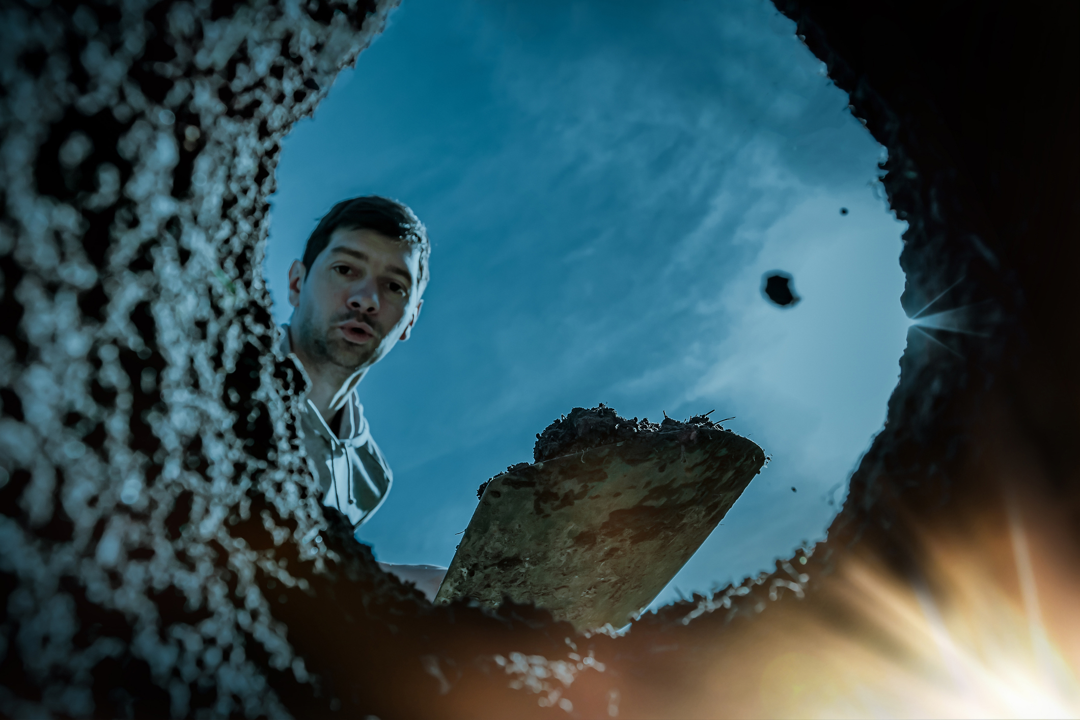 Man with a surprised expression, holding a spade, peers down into a hole with bright light shining from the bottom. Sky is visible overhead