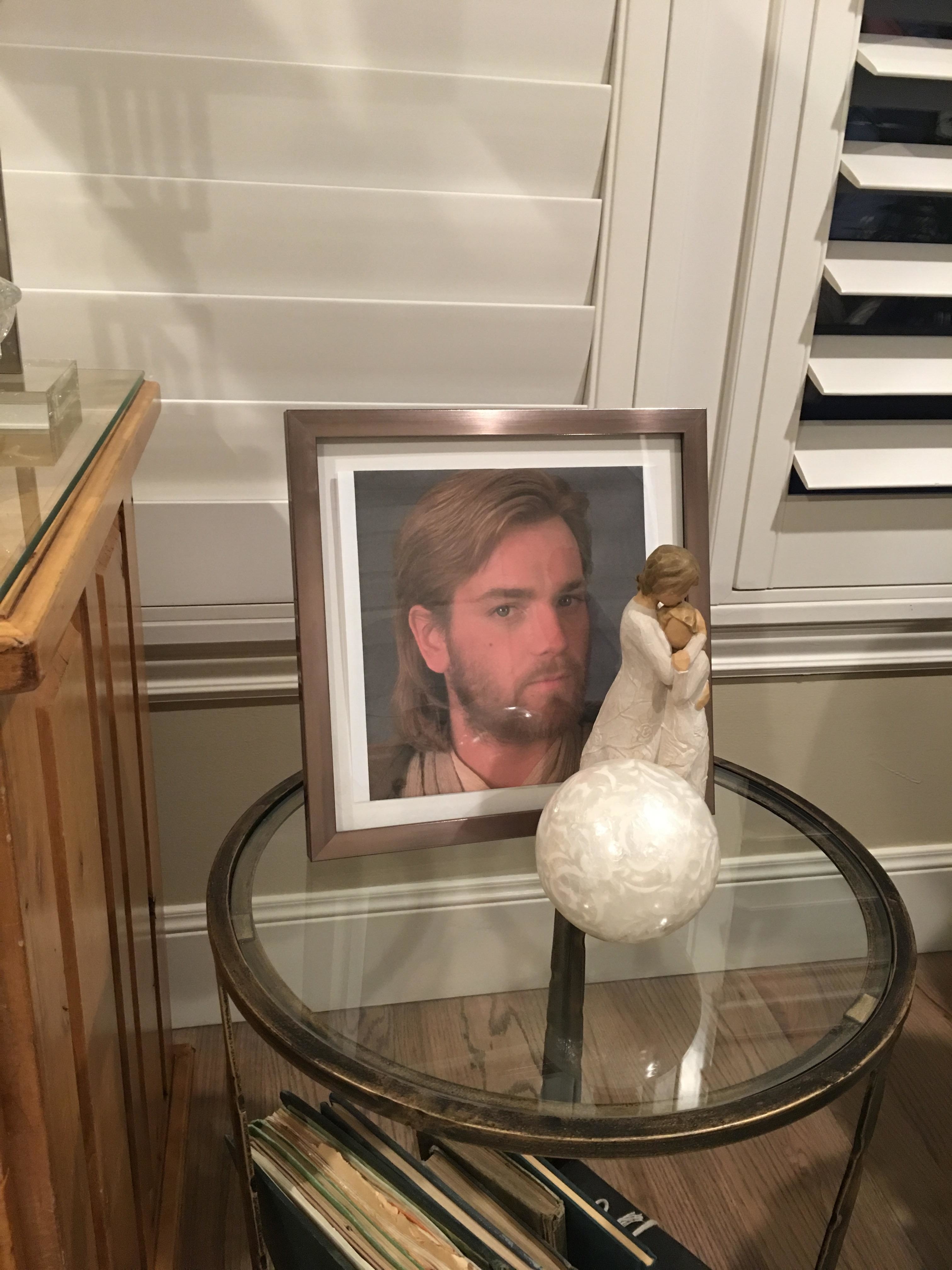 A framed photo of a man with long hair and a beard is on a small round table next to a figurine of a mother holding a child