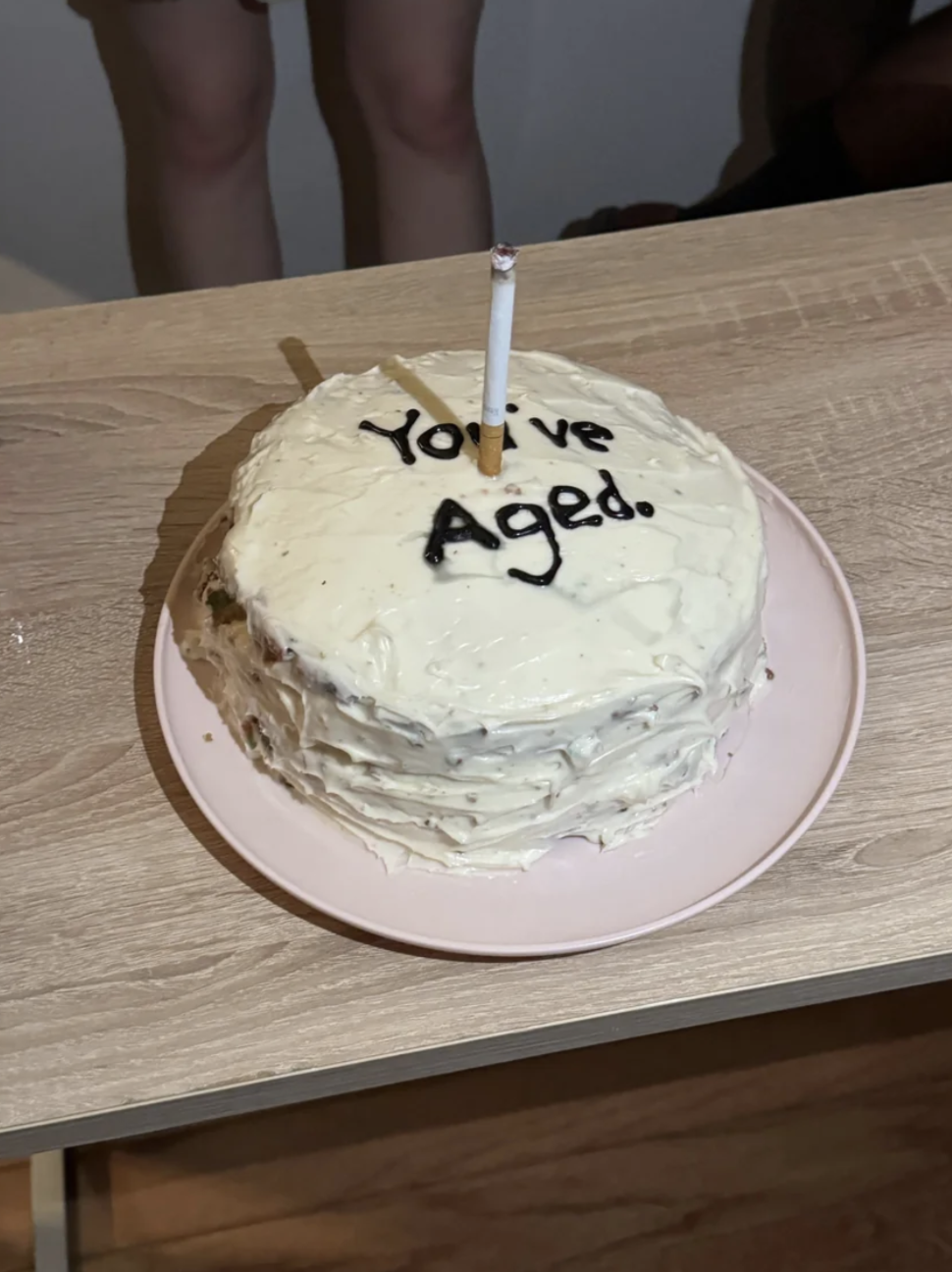 A round cake with white frosting has a single lit candle and the text "You've Aged." written on top in black frosting. It is on a light-colored plate
