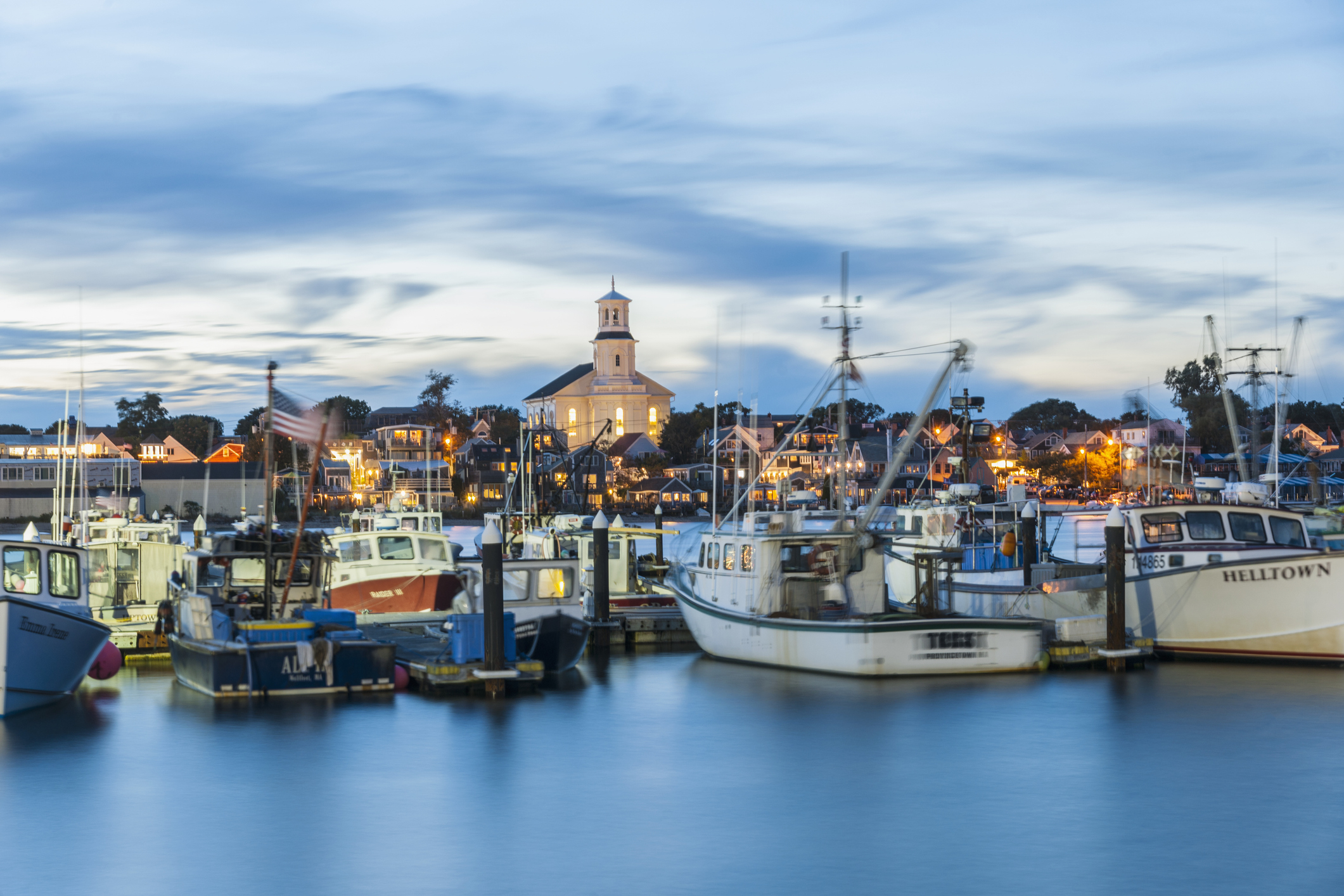 A peaceful harbor scene with several fishing boats docked in calm water, a town with lit buildings and a church steeple is visible in the background