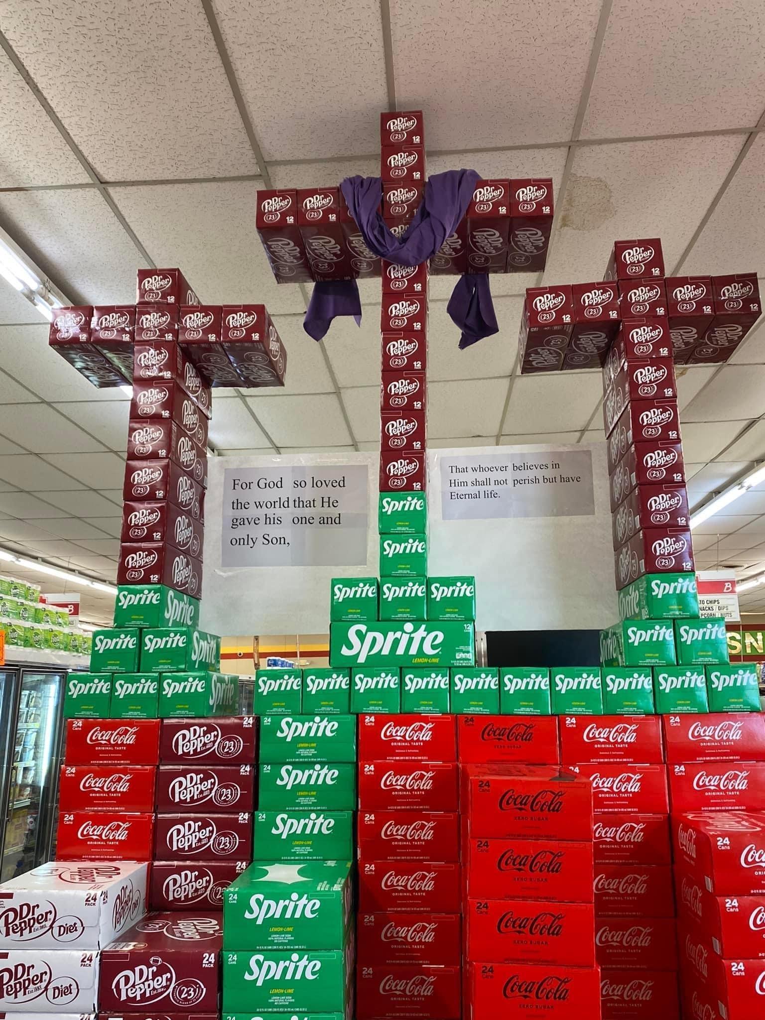 Grocery store display with soda boxes arranged as three crosses. The central cross is adorned with a cloth. Bible verses are displayed above the crosses