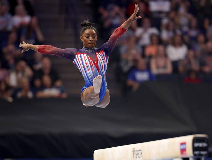 Simone Biles performs a split leap mid-air on the balance beam during a gymnastics competition, wearing a leotard with a red, white, and blue pattern