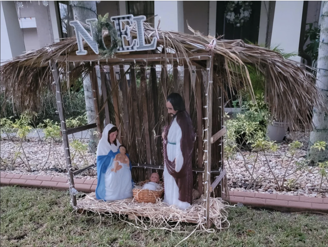 Nativity scene with figures of Mary, Joseph, and baby Jesus in a manger under a thatched roof. The word "NOEL" is displayed above the scene