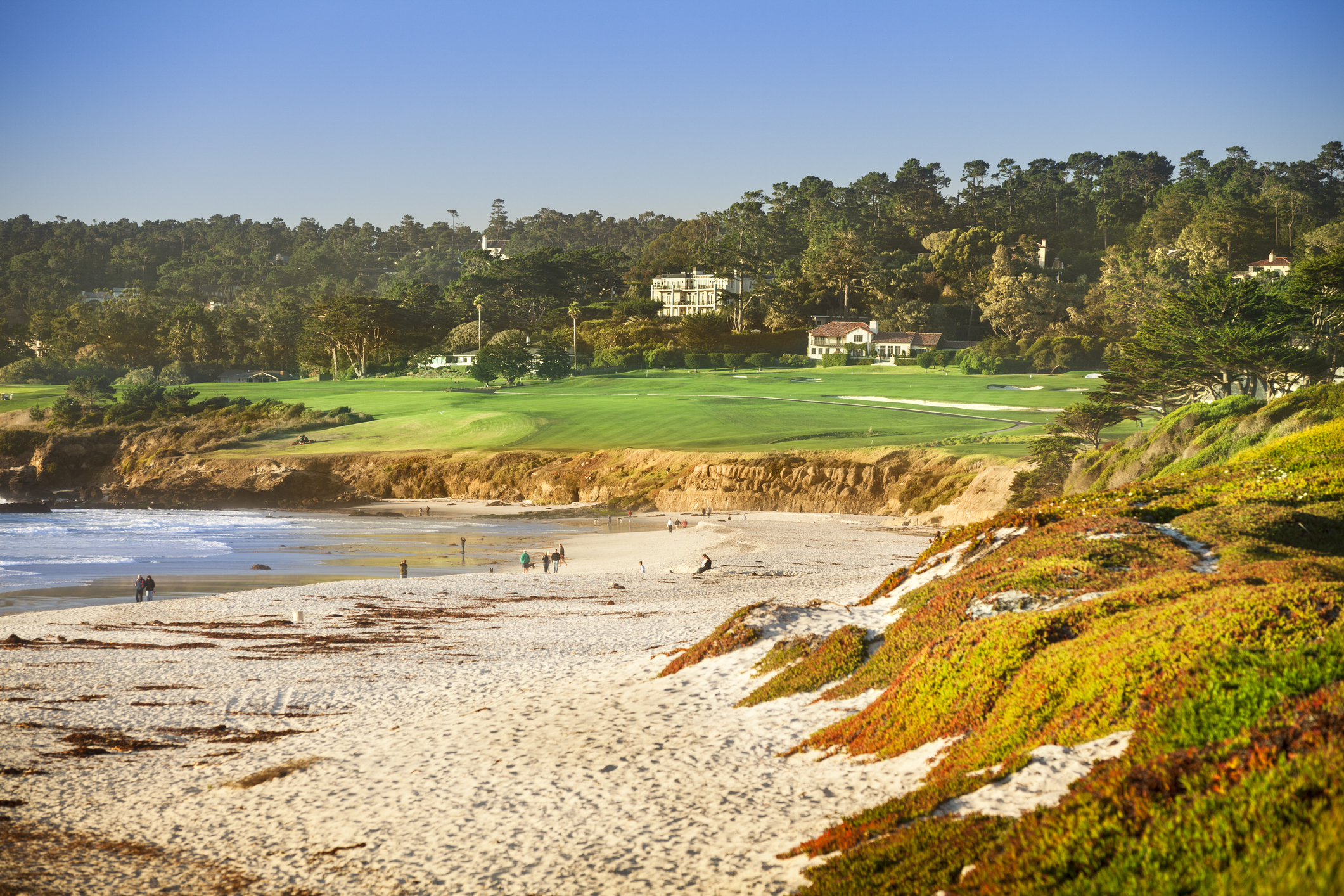 A scenic beach with waves crashing on the shore, backed by a lush golf course, buildings, and trees on a hillside under a clear sky