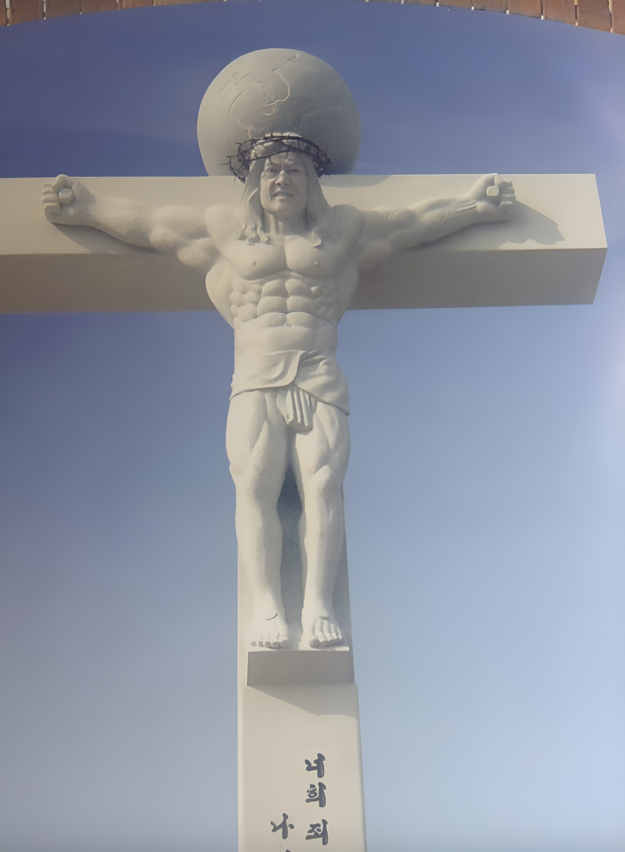 A statue of a muscular man wearing a headpiece, resembling a crucifixion scene, with scripting along the bottom of the cross