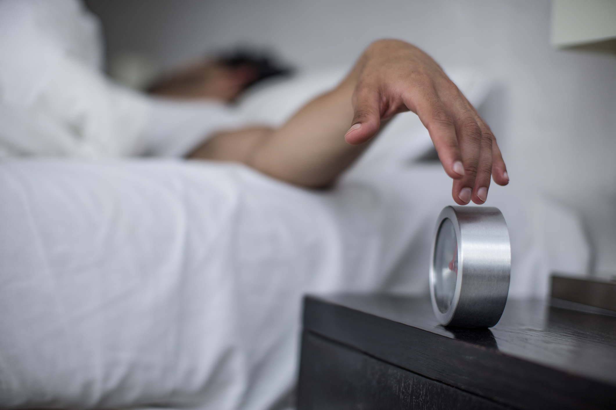 A person lying in bed reaches out to turn off a silver alarm clock on a bedside table