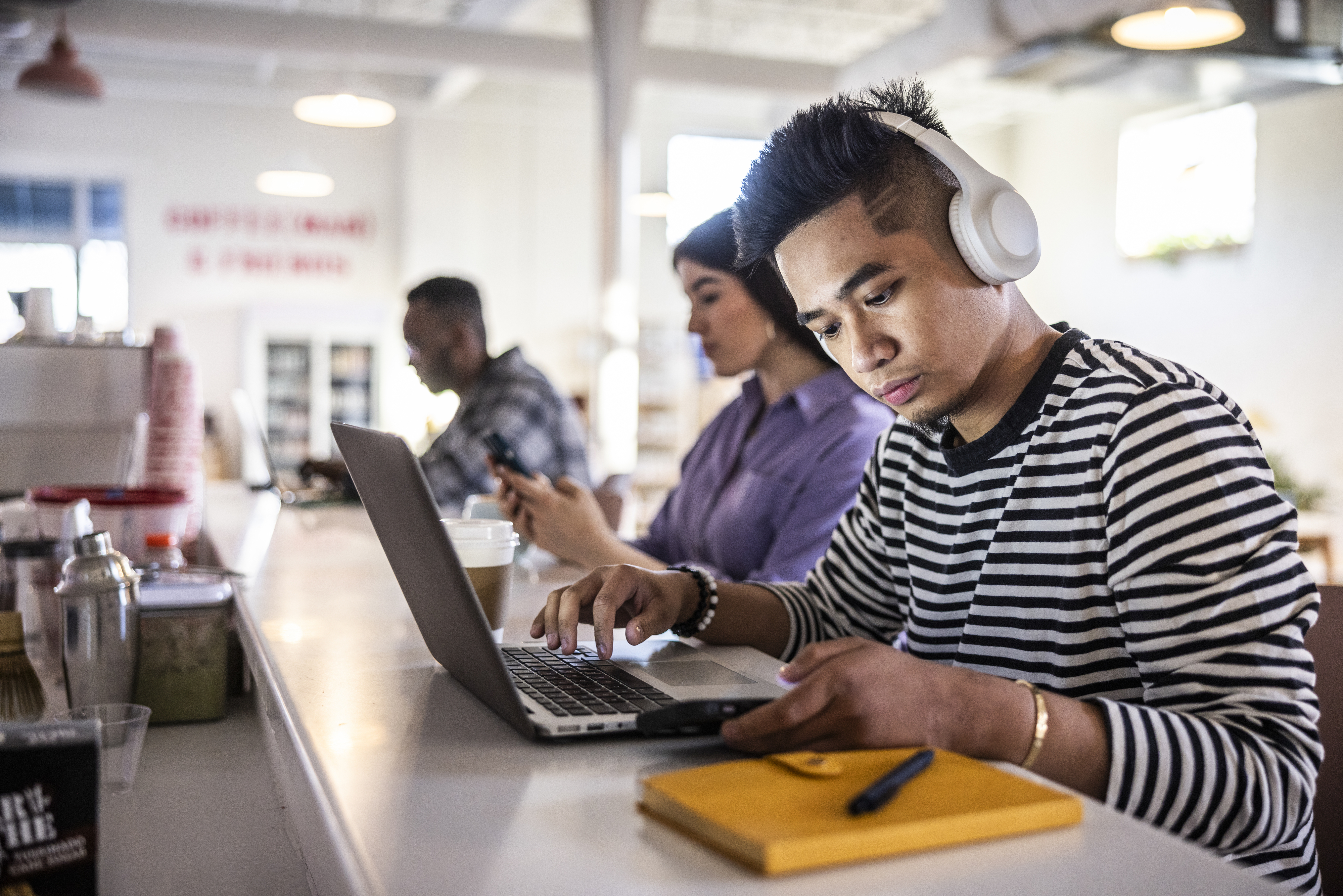 Three people work on laptops and phones at a shared desk in a modern cafe. One person listens with headphones, wearing a striped shirt. Others are in casual attire