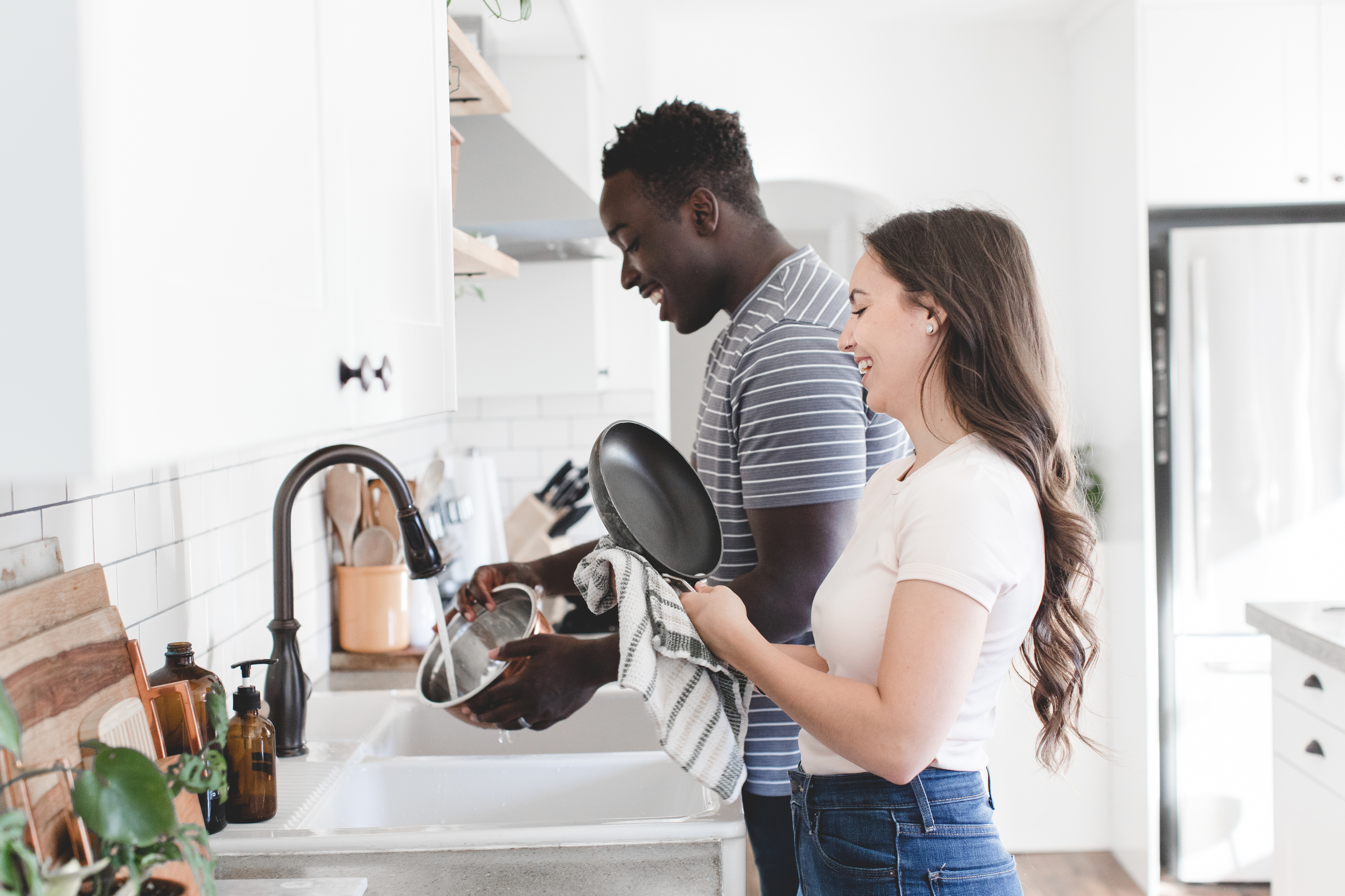 Two people, one holding a pan and the other washing dishes, smile and work together in a modern kitchen