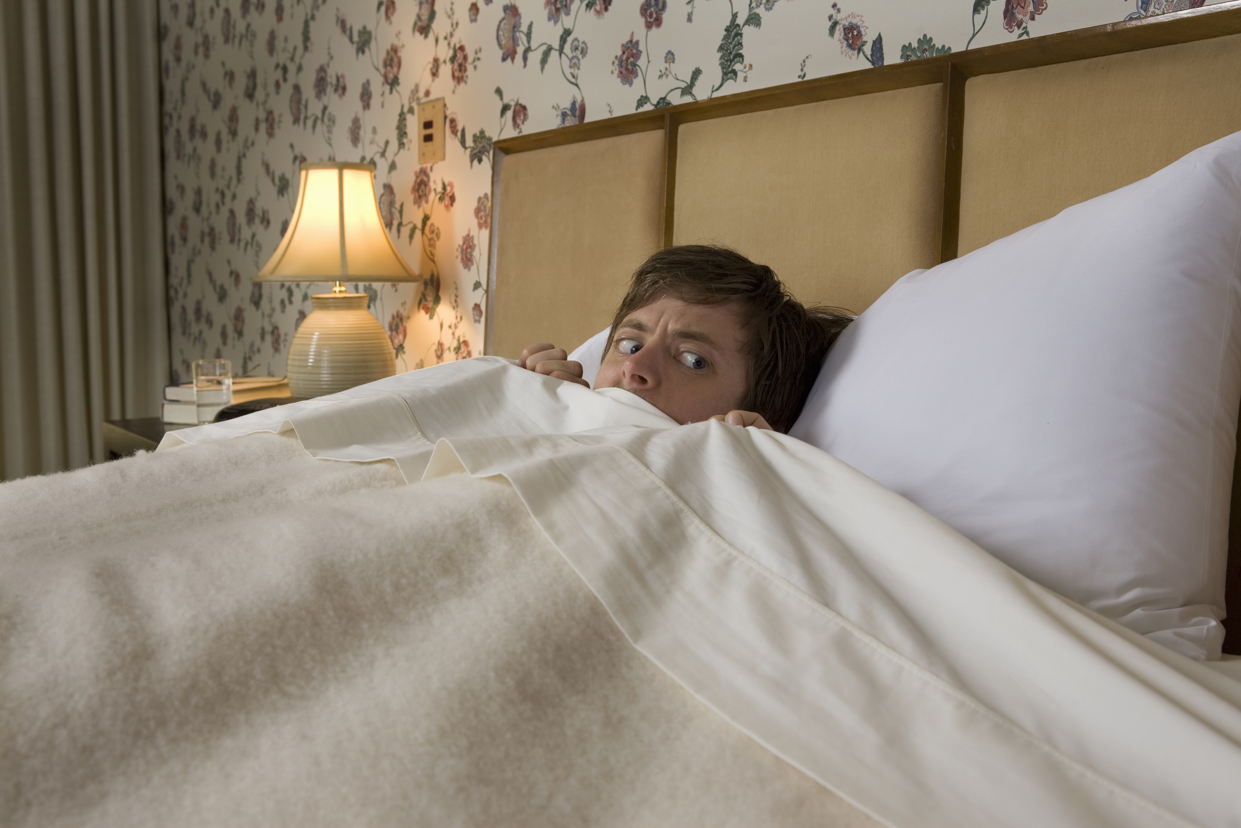 Man lying in bed, looking scared, with the blanket pulled up to their chin