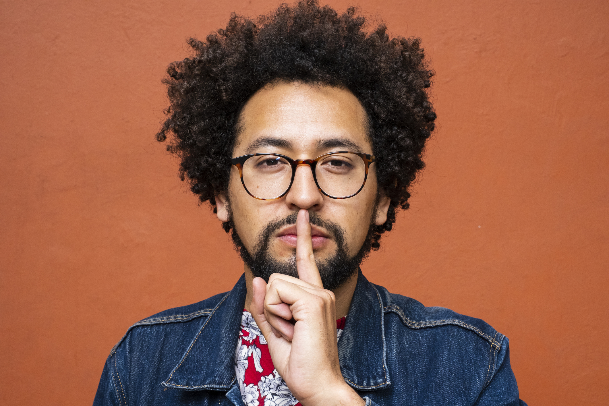 Person with curly hair and glasses making a shushing gesture with a serious expression, wearing a denim jacket and patterned shirt