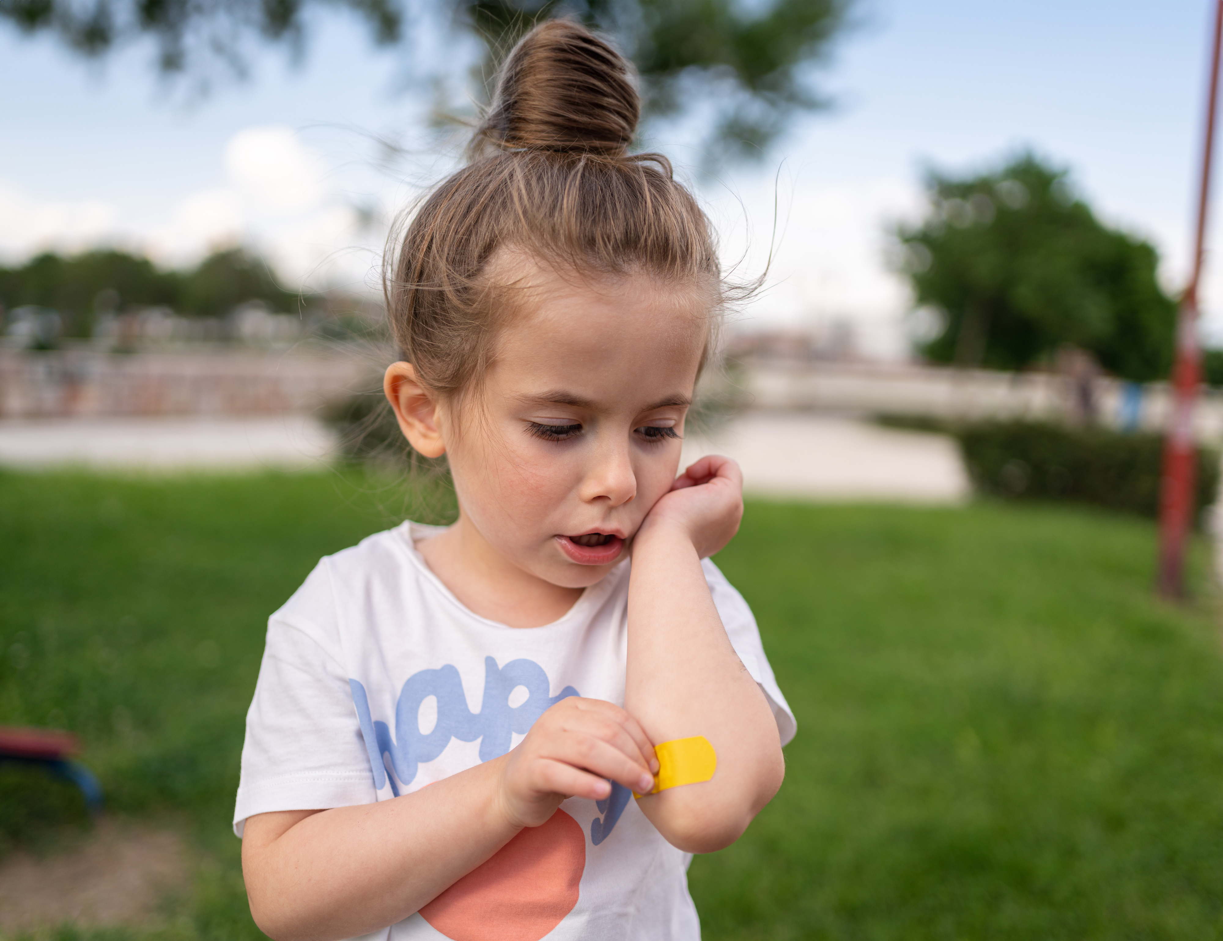 A young girl inspects a bandage on her arm while standing outside in a park