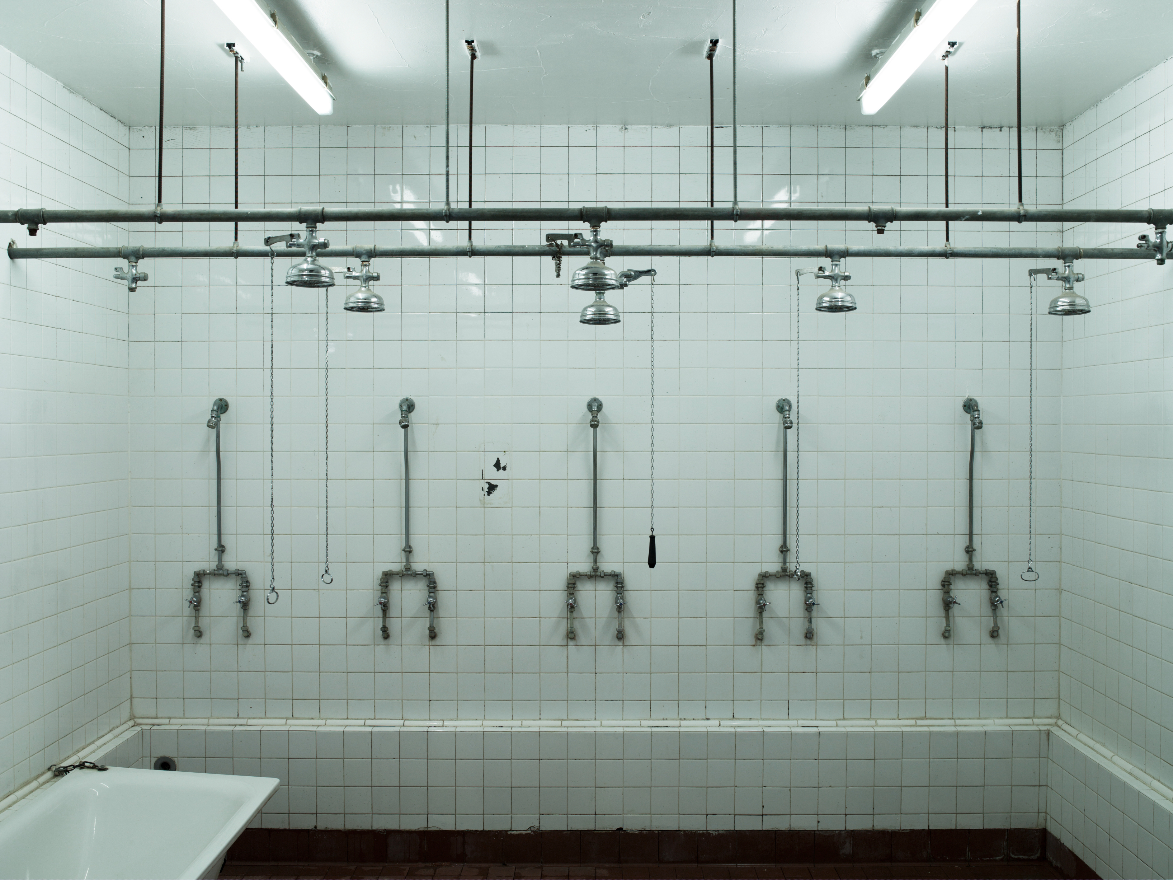 A white-tiled shower room with six shower heads mounted on the wall, each with separate faucets and pipes. The room appears to be industrial or institutional