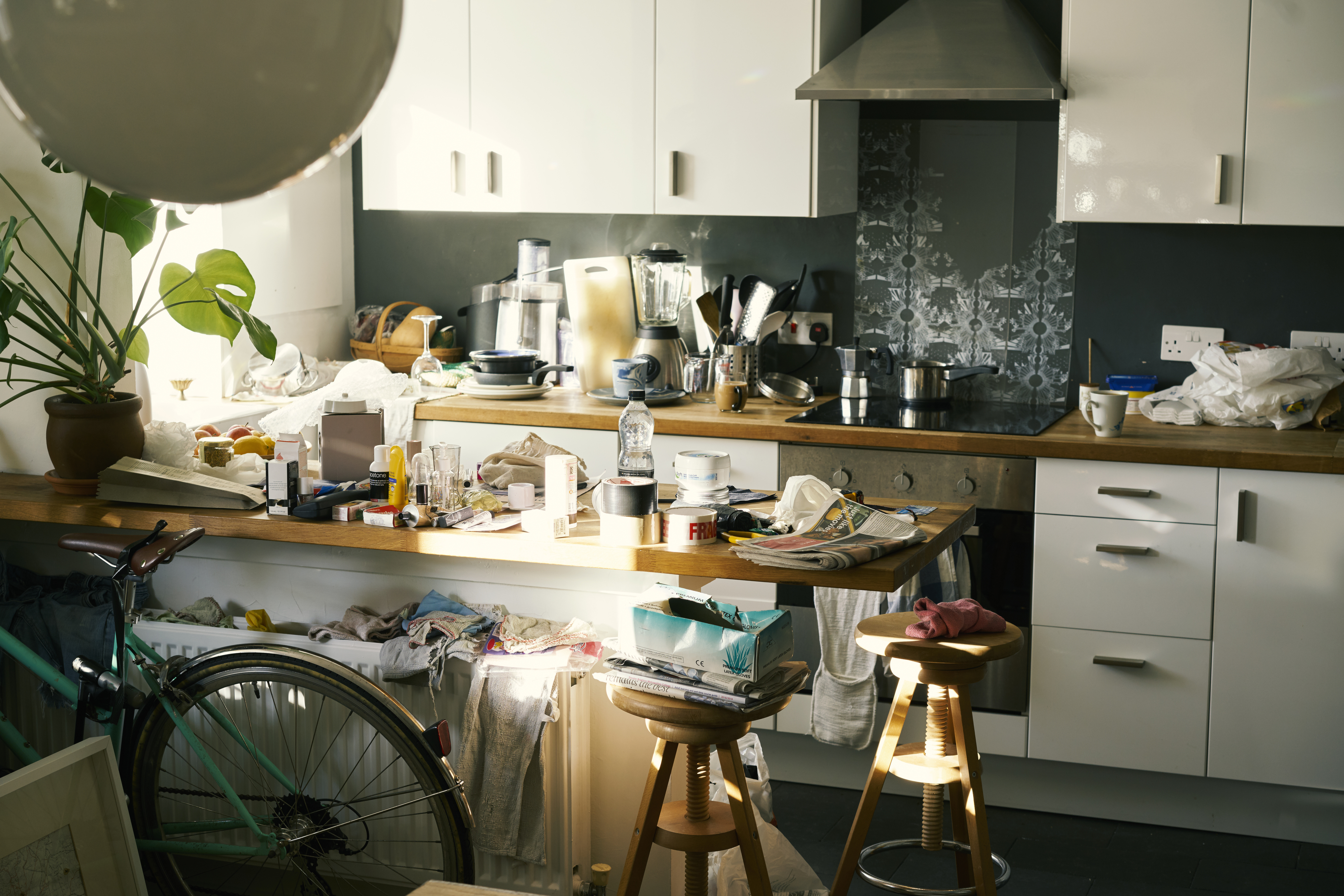 A cluttered kitchen with multiple items scattered on the counter, including utensils, appliances, and a bicycle leaning against the counter