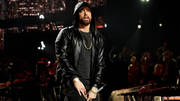 Eminem performs on stage in a black leather jacket, hoodie, and gold chains, holding a microphone