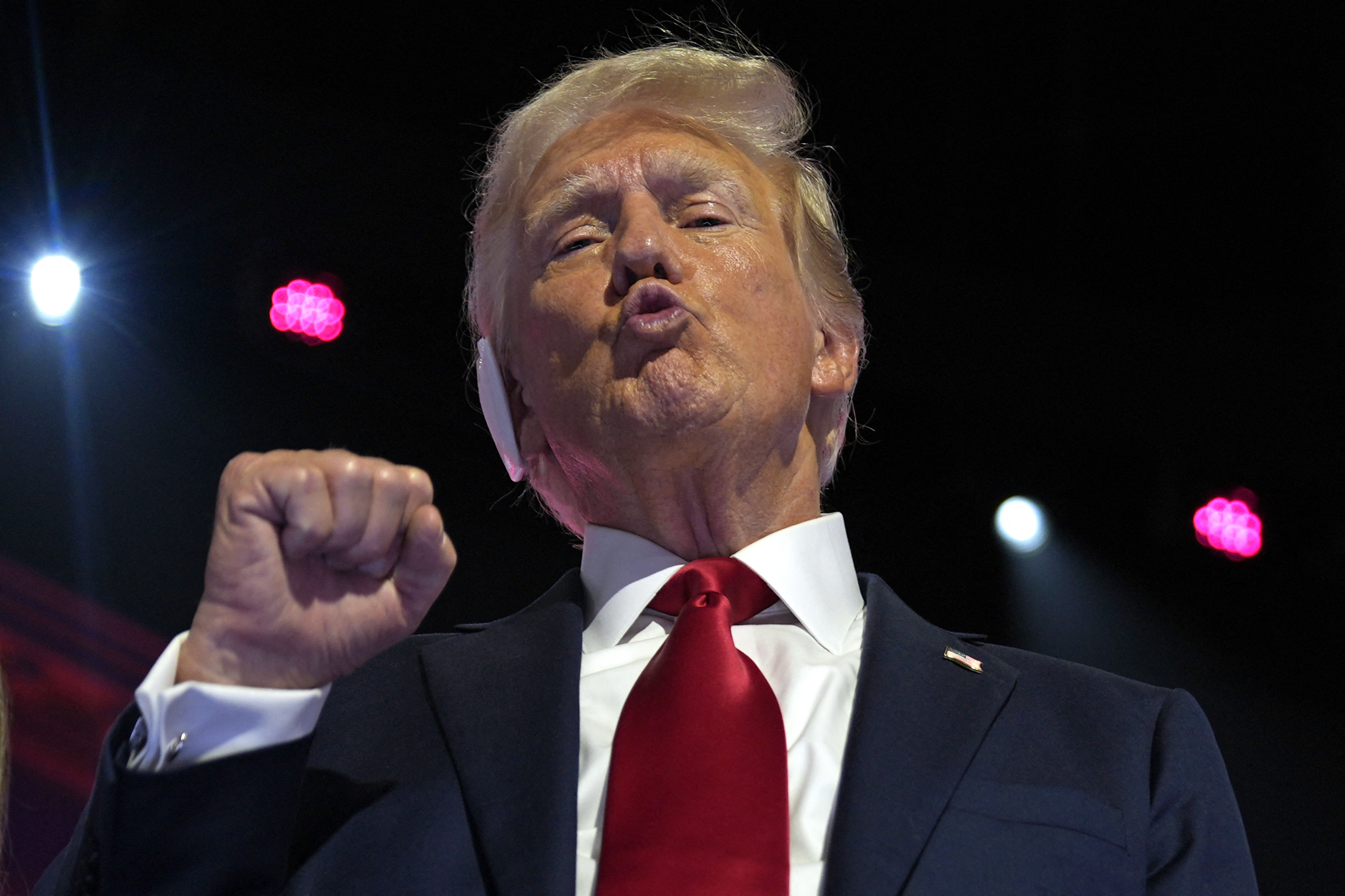 Donald Trump, wearing a dark suit and red tie, gestures with a raised fist, standing on a stage with bright lights in the background