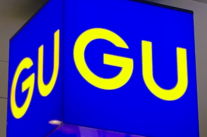 Bright blue sign with “GU” in large yellow letters hanging from a ceiling with recessed lighting