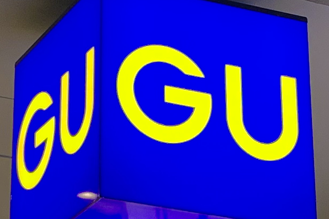 Bright blue sign with "GU" in large yellow letters hanging from a ceiling with recessed lighting