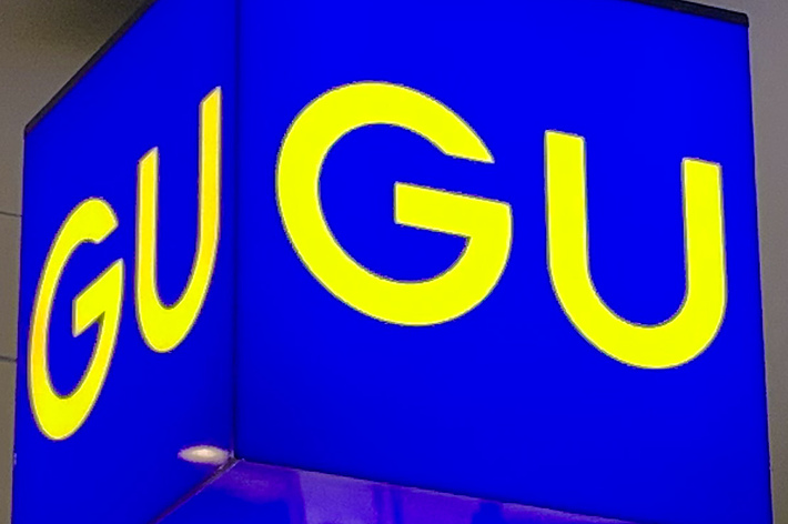 A blue, cube-shaped sign with the letters &ldquo;GU&rdquo; in yellow on each visible face. The sign is illuminated and hanging indoors
