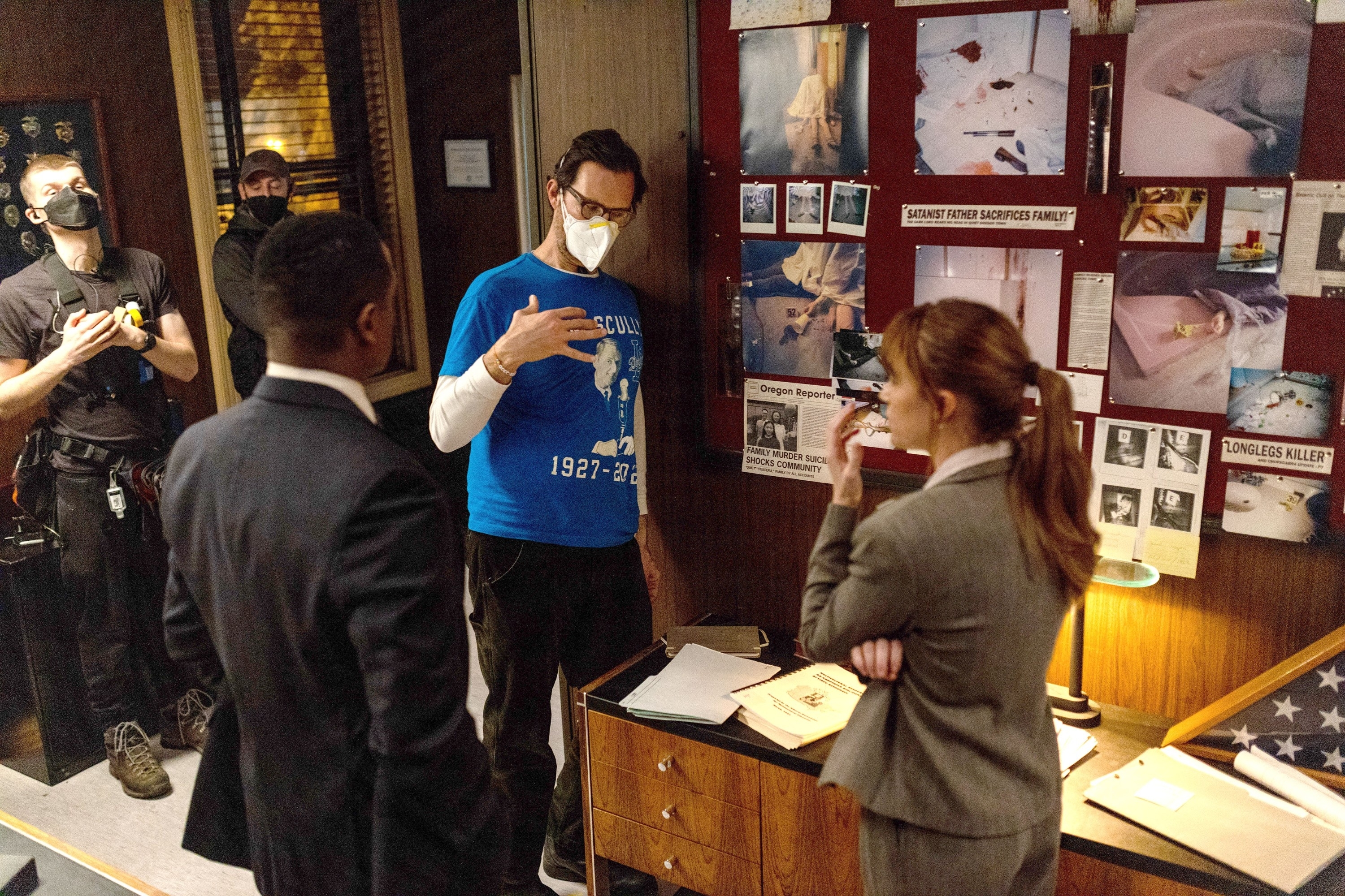 Four crew members are discussing a scene on a crime drama set, examining a board with evidence photos
