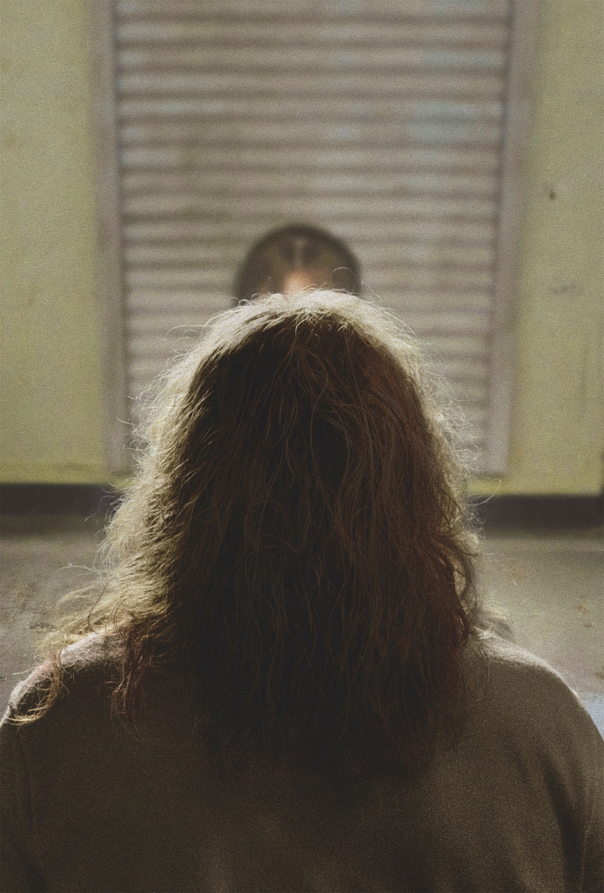 Person with long, wavy hair stands facing a weathered, partially open door with a face barely visible behind it in a scene from a TV show or movie