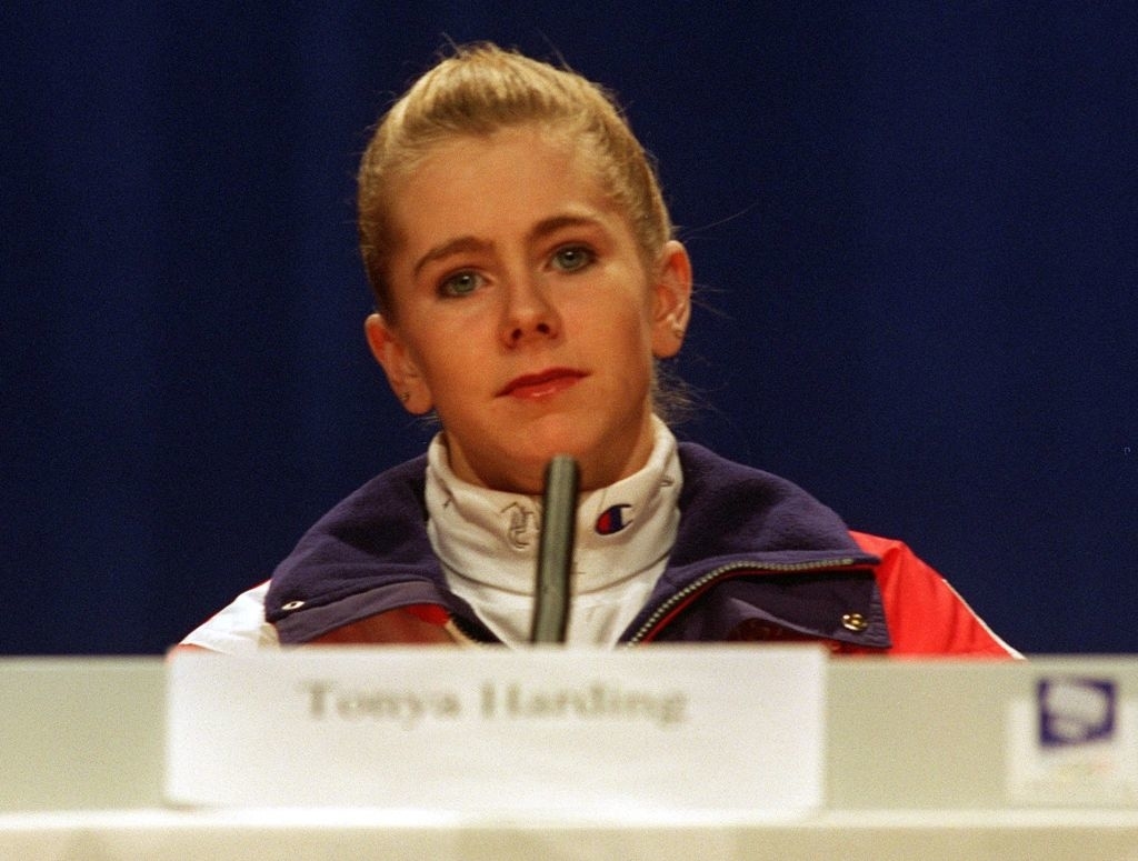 Tonya Harding sits at a press conference, wearing a sporty jacket with a white turtleneck