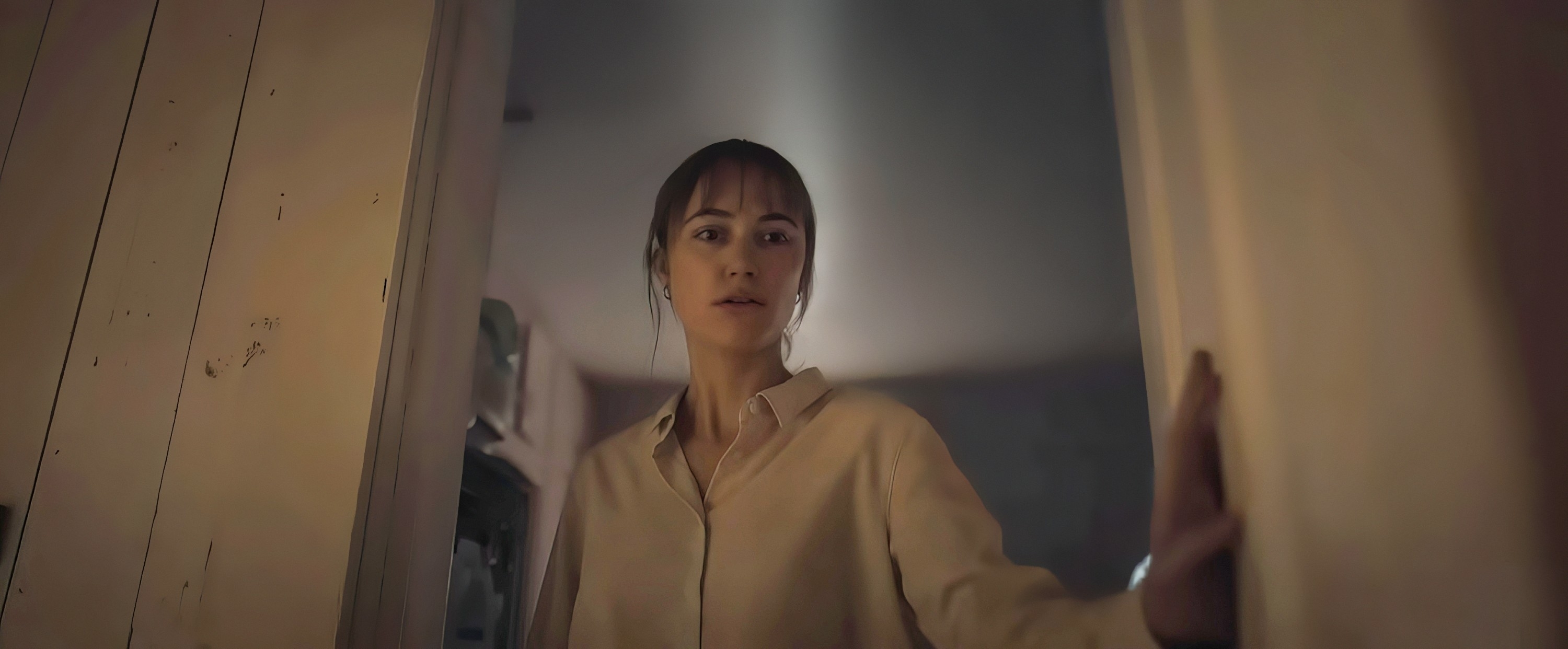 Actress Sydney Sweeney cautiously peeks through an open door, dressed casually in a light shirt