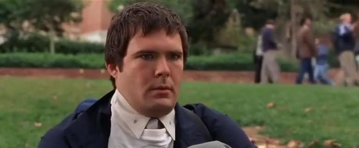 Actor from a movie scene sits on grass with a serious expression, wearing a jacket and an untied necktie, with blurred people walking in the background