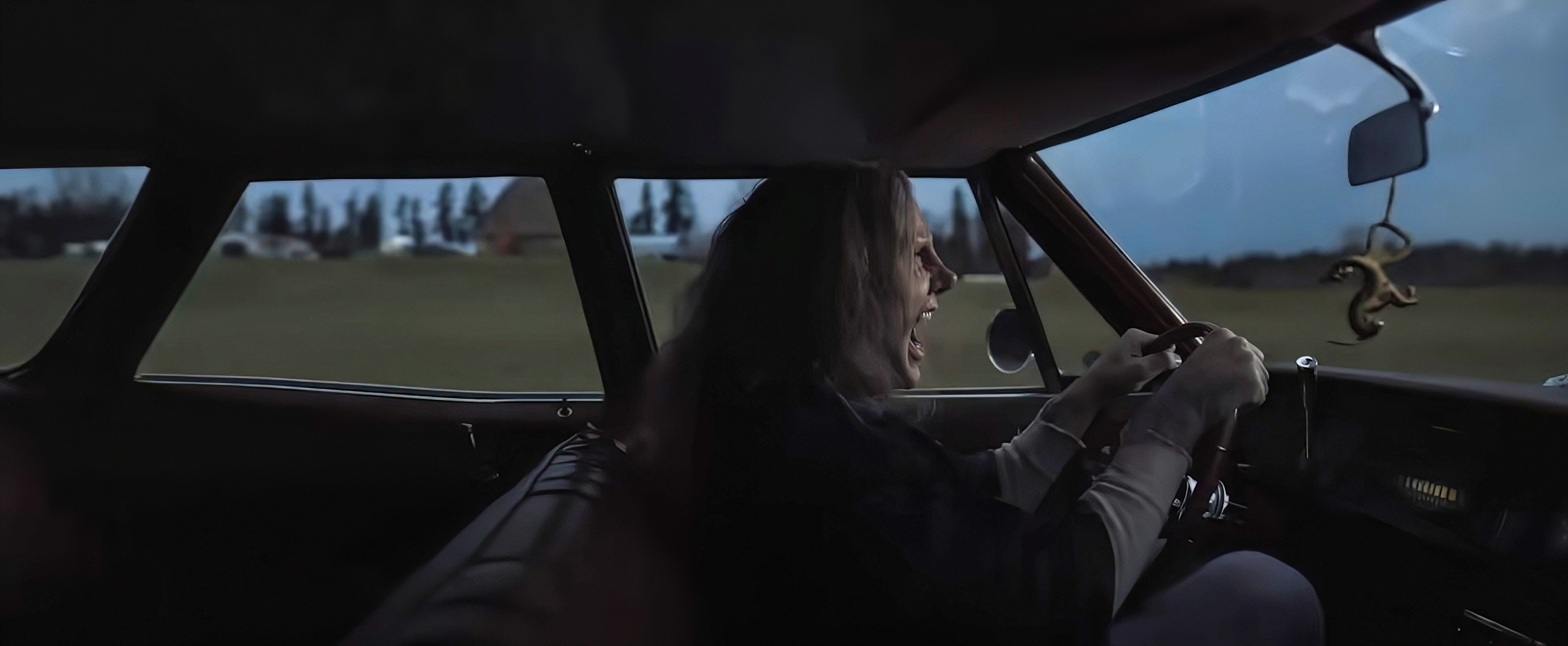 Man passionately singing while driving an old car in a rural area with trees and houses in the background