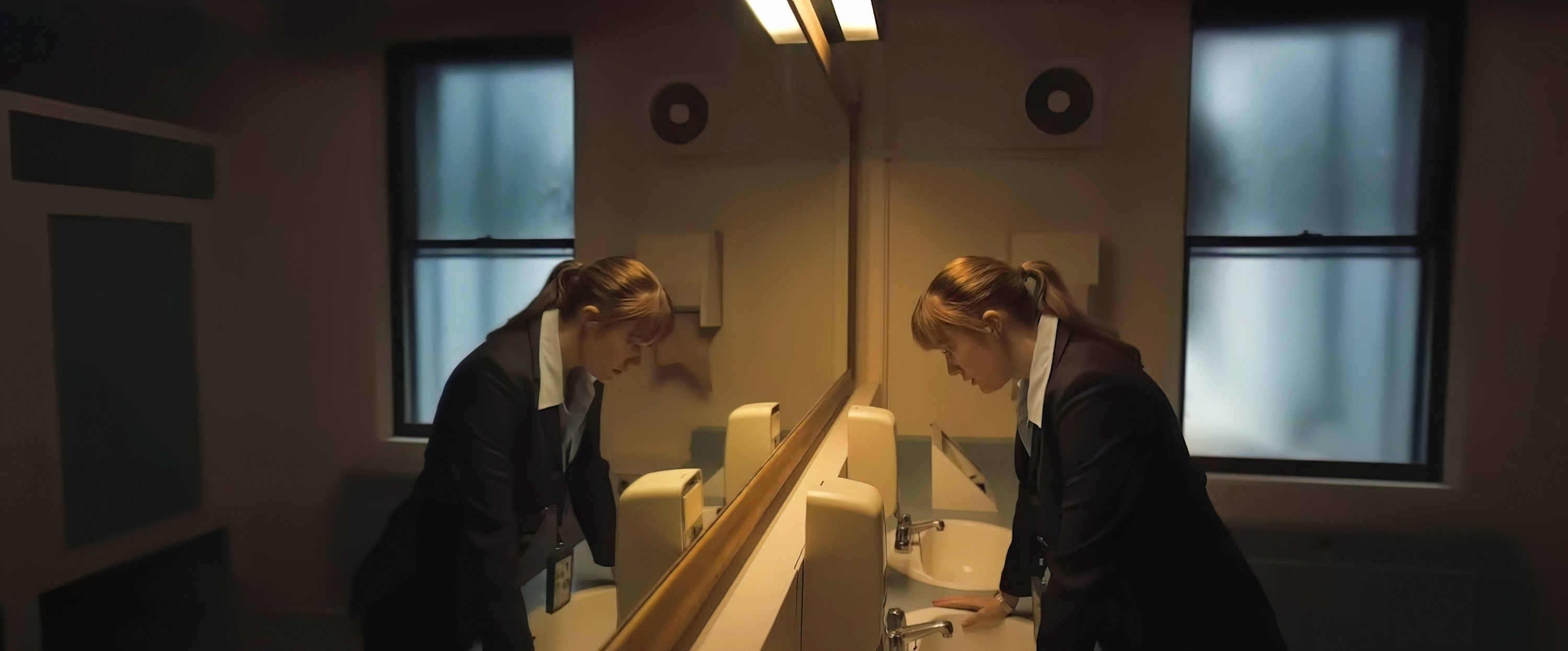 Jodie Comer, in a scene from the production set of TV Show "Killing Eve," wears a dark blazer with a white shirt, standing by a bathroom sink and looking down