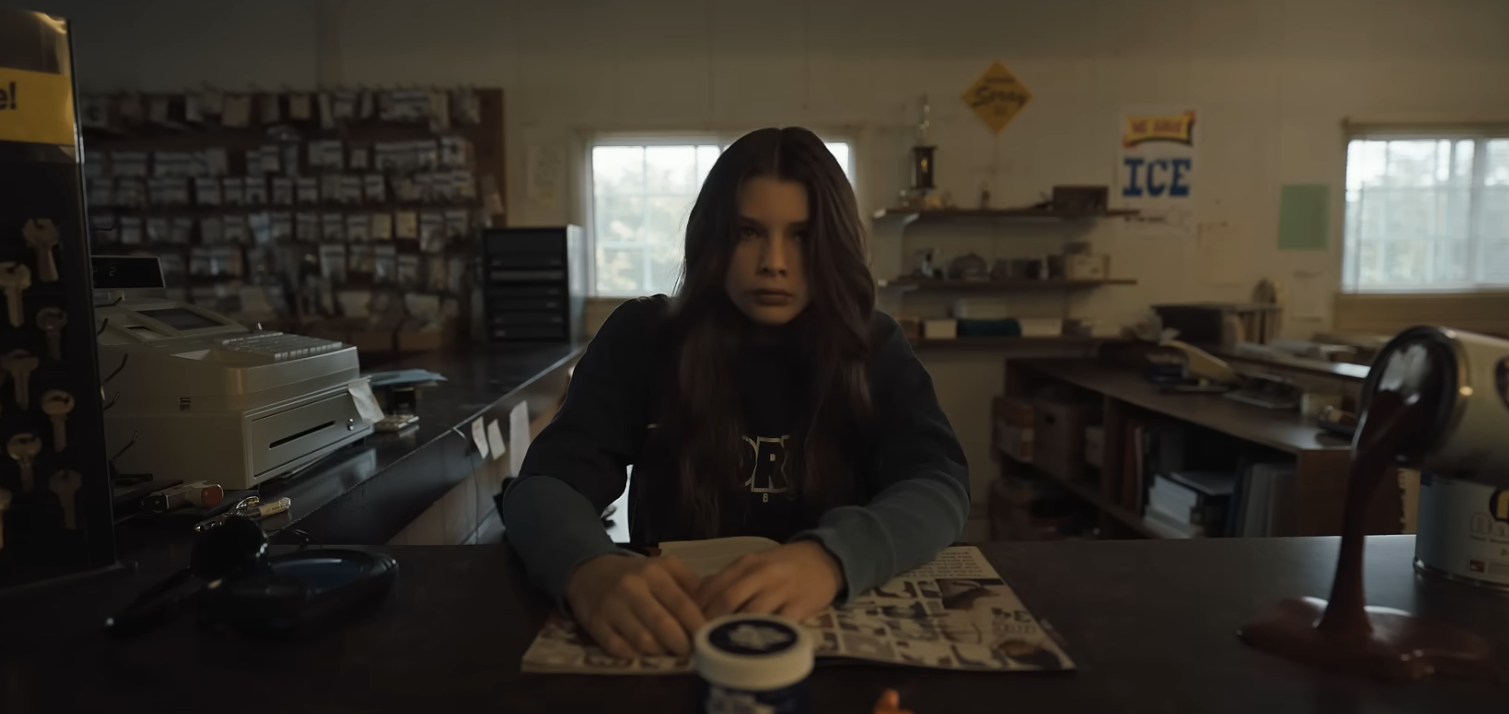 Young girl with long hair intensely reading a newspaper at the counter of what appears to be a hardware store