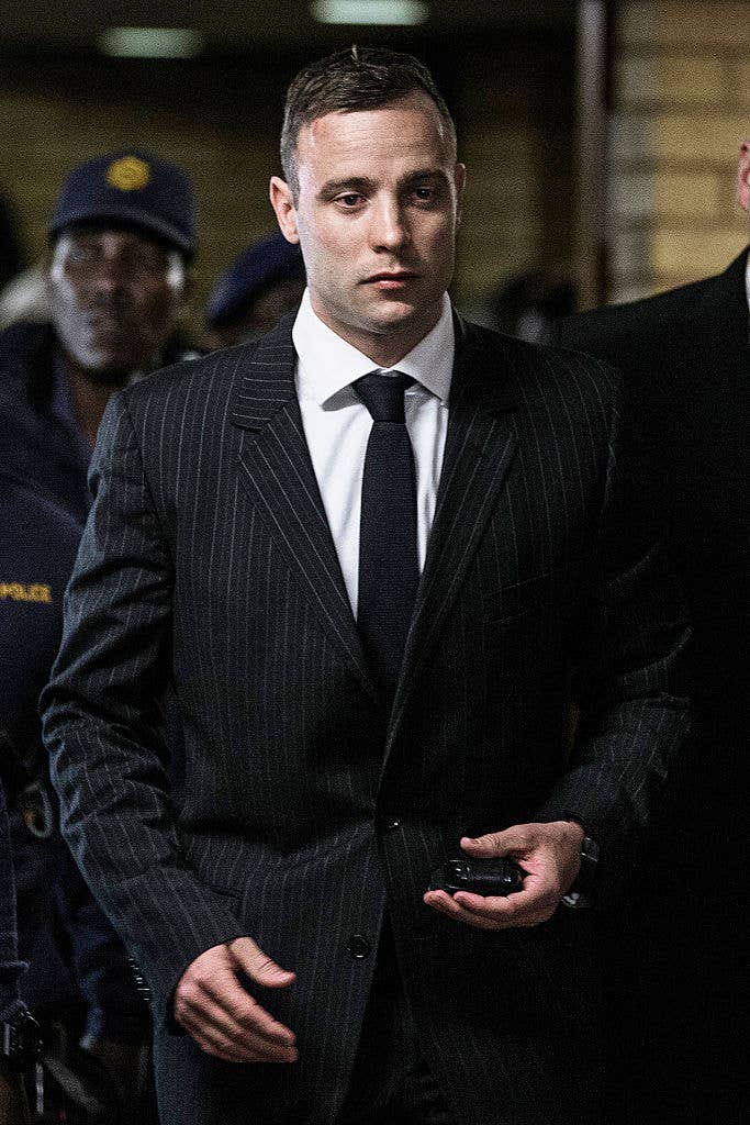 Oscar Pistorius, in a pinstripe suit with a white shirt and dark tie, walks with police officers in the background