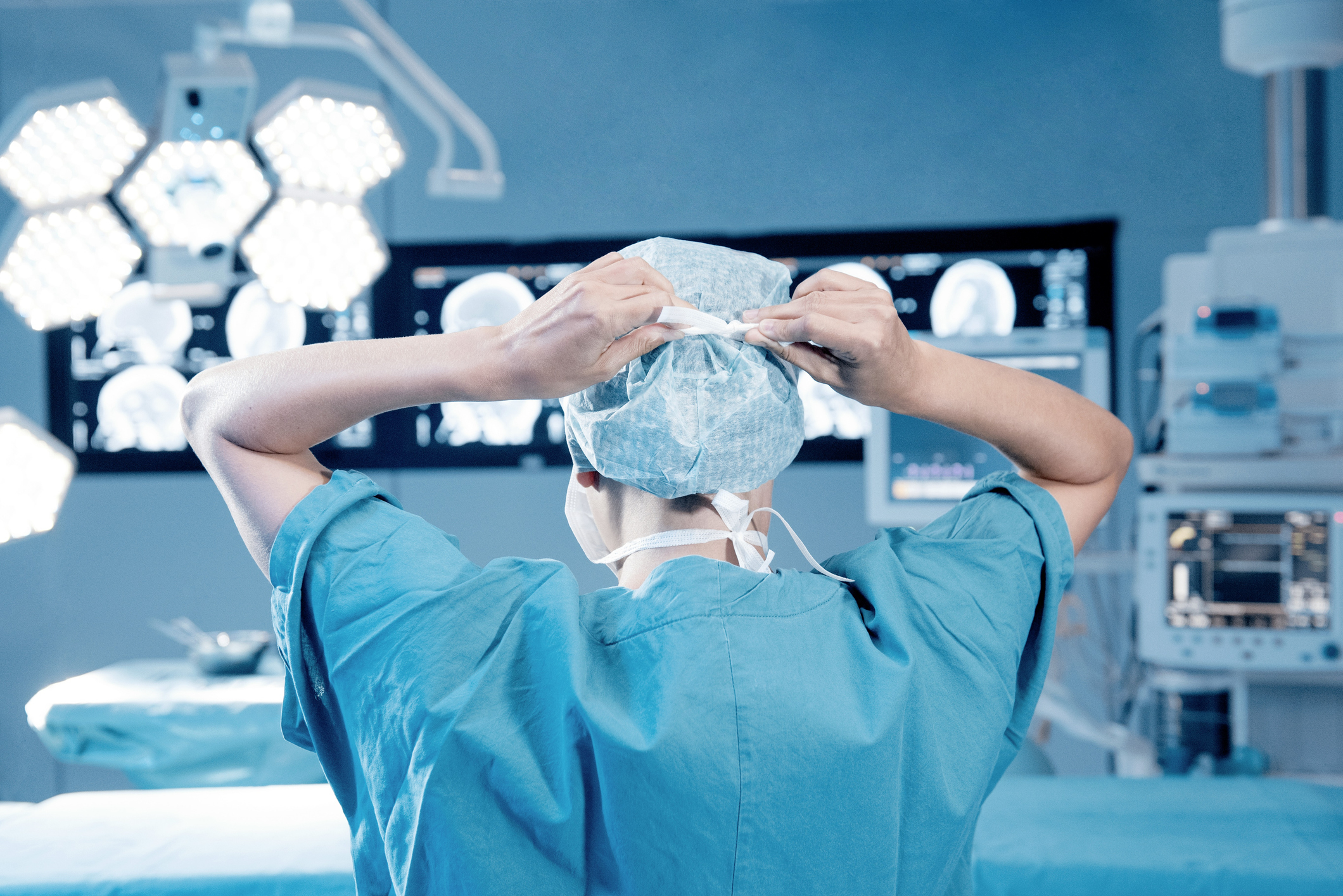 A surgeon in an operating room adjusts their cap, facing away from the camera, with medical equipment and monitors displaying brain scans in the background