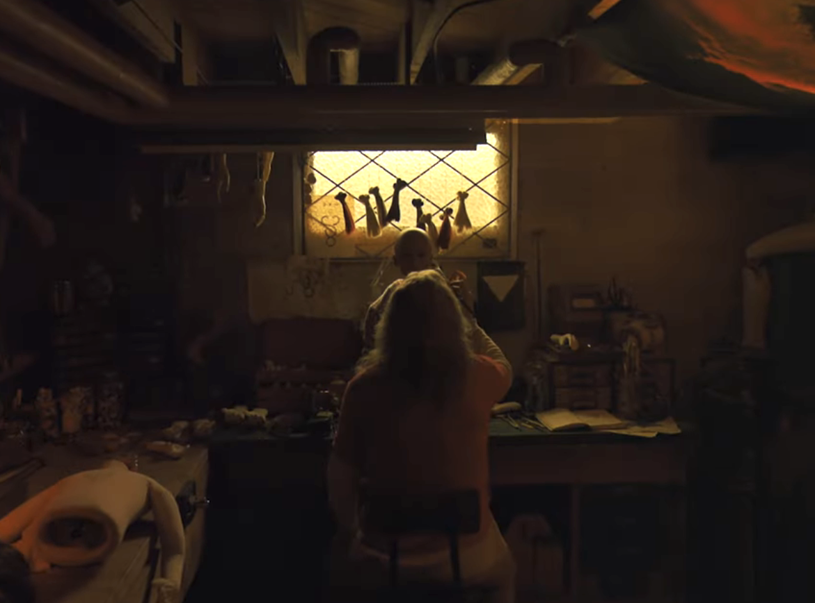 A person sits at a table in a dimly lit room, facing a window with letters taped to it. Various objects and paper clutter the table and surrounding area