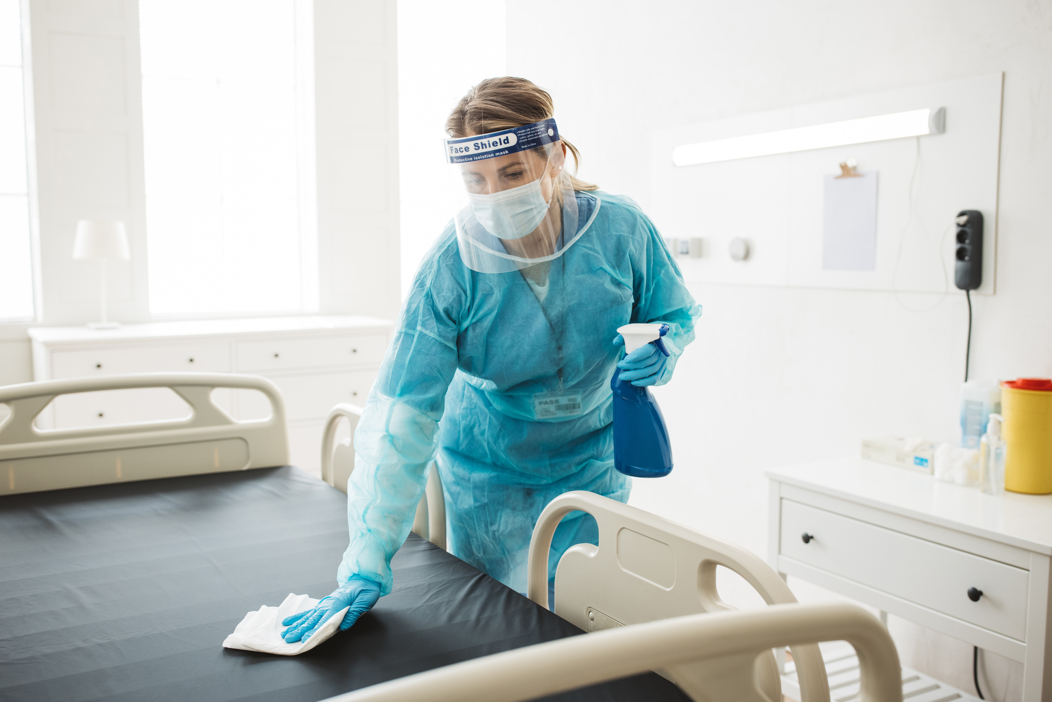 A healthcare worker in protective gear, including a face shield, mask, gloves, and gown, cleans a hospital bed with a spray bottle and cloth