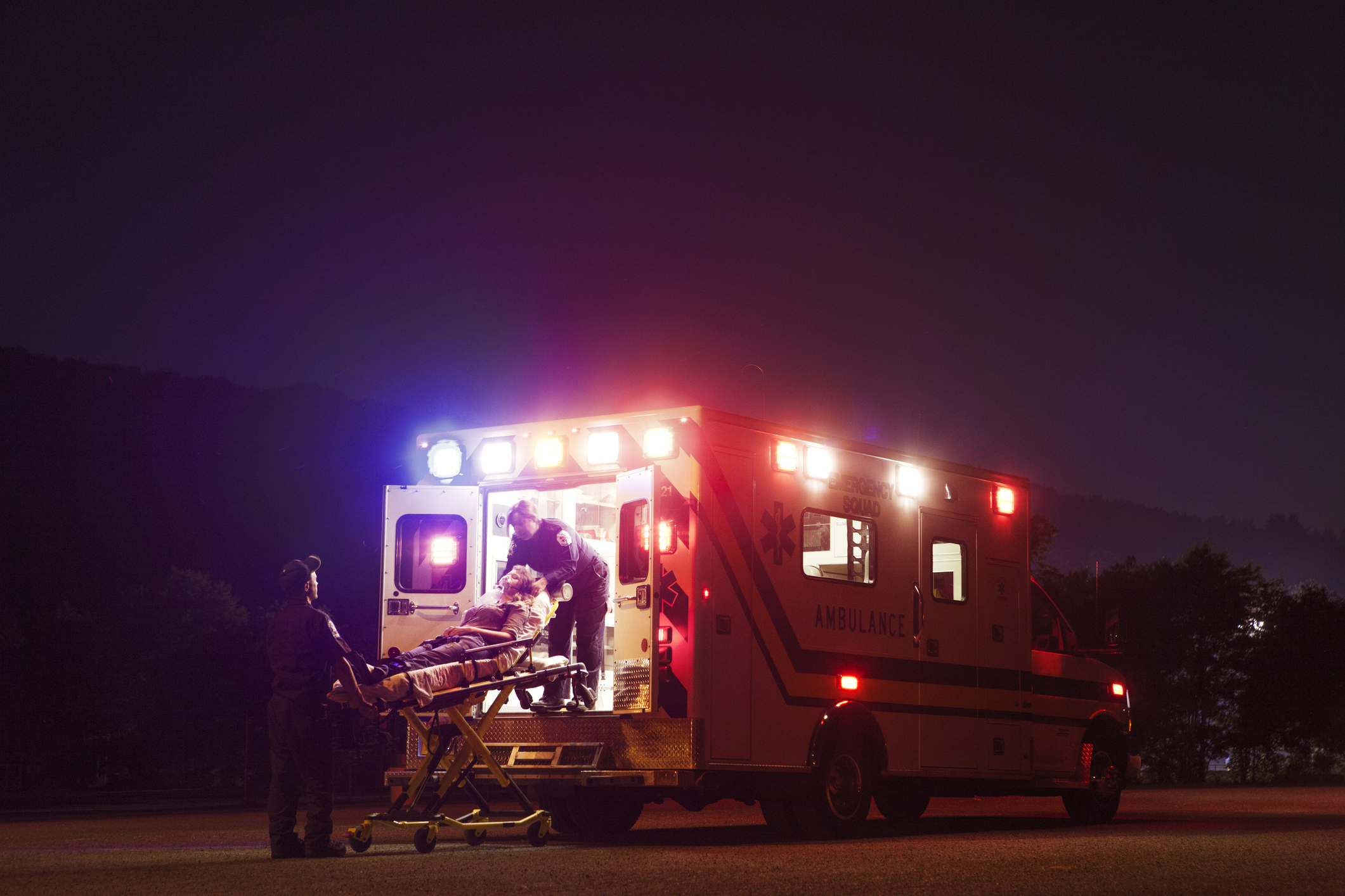 A patient is being loaded into an ambulance at night by emergency responders. The ambulance lights are flashing