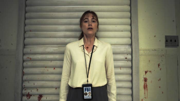 Gillian Anderson stands in front of a metal shutter, wearing an FBI badge and a white blouse, in a scene from a TV show