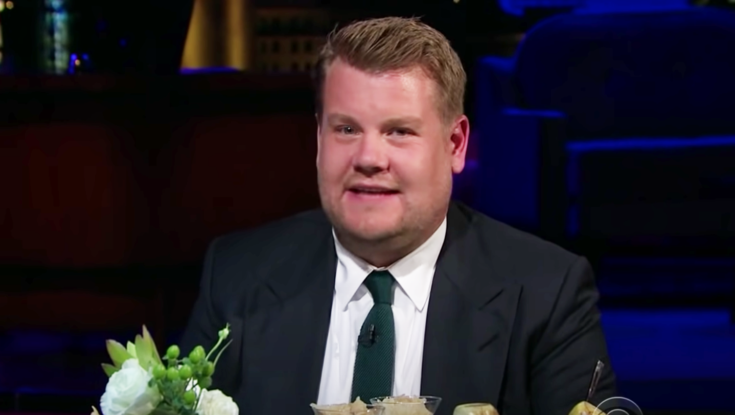 James Corden sitting at a table during a talk show, dressed in a suit and tie with flowers and food on the table in front of him