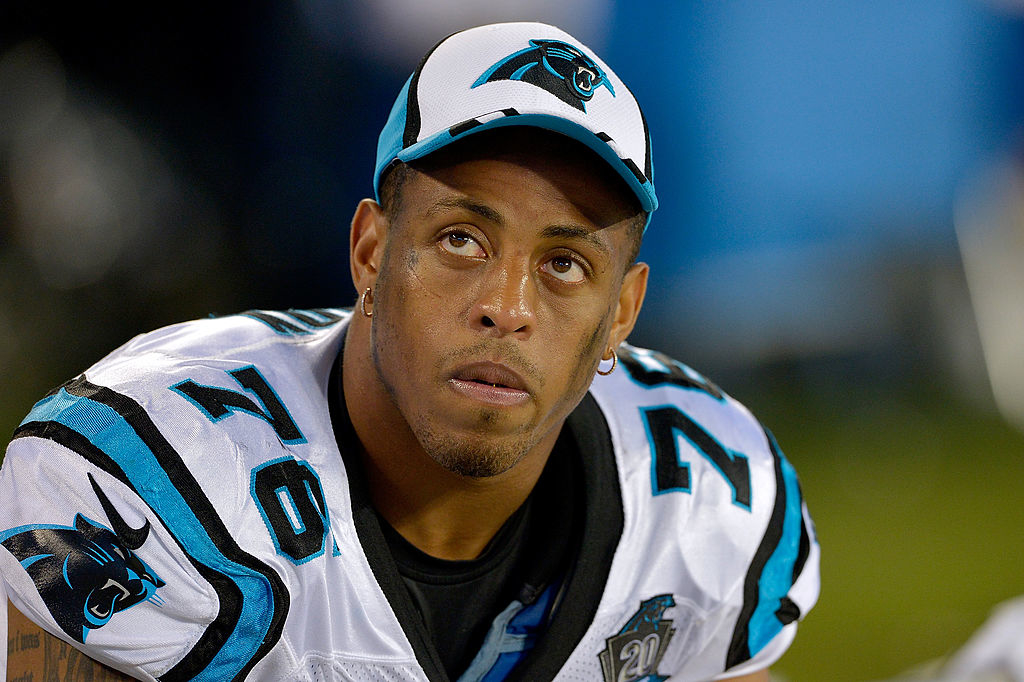 Greg Hardy, in a Carolina Panthers football uniform, looks up while sitting on the sidelines during a game, wearing a cap