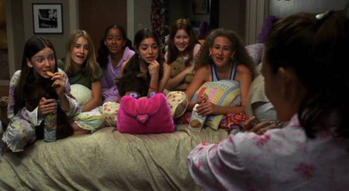 Seven girls in pajamas gather on a bed with snacks, listening to the girl facing them. They appear to be having a sleepover